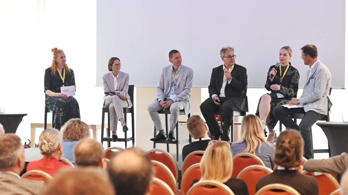 Sechs Personen sitzen nebeneinander auf einer Bühne mit Publikum vor sich.