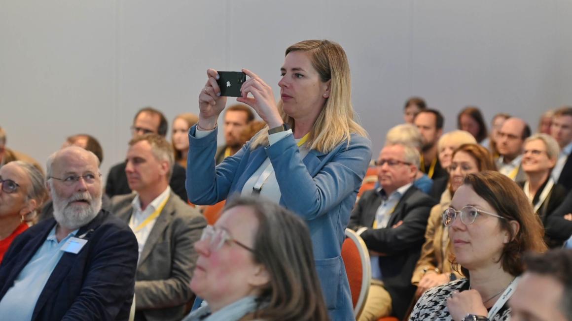Eine stehende Frau im Publikum fotografiert mit einem Handy in beiden Händen.